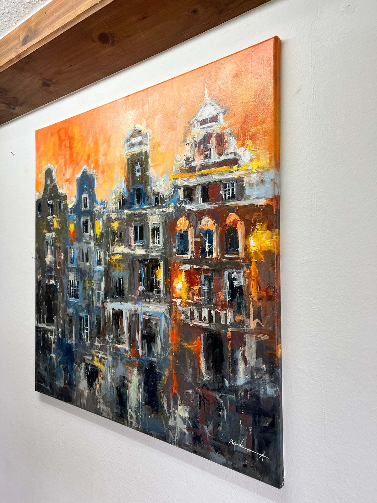 An expressive canvas painting of amsterdam city buildings at dusk, shown hanging on a white wall under a wooden ceiling beam. The artwork features warm orange and yellow light reflections against dark blue, gray, and black architectural forms.