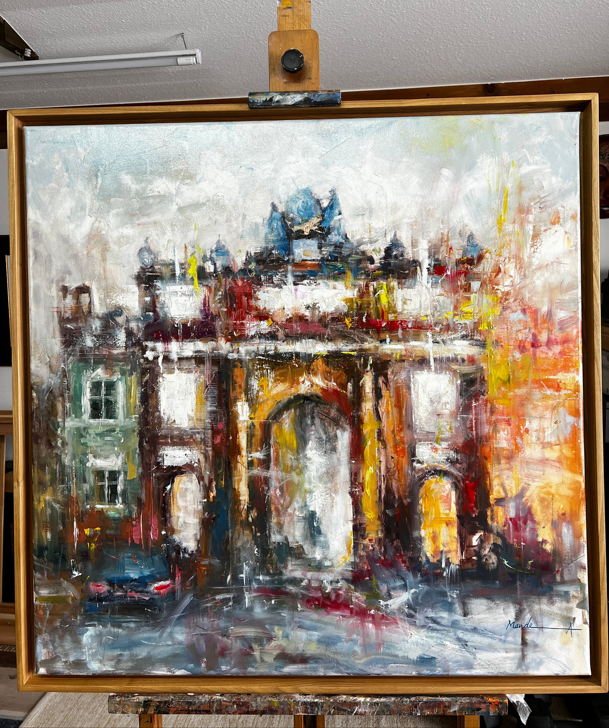 An impressionistic oil painting of a historic city gate or archway, displayed on an easel. The artwork features thick impasto brushstrokes in warm tones of orange, yellow, red, and brown, contrasted with cool gray, white, and light blue tones in the sky and street reflections.