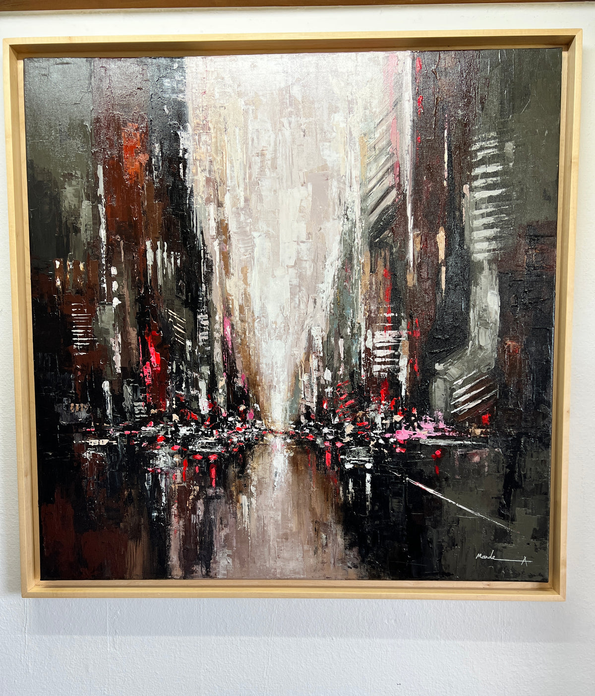 An abstract framed painting depicting a city street scene with tall dark buildings, a bright central vanishing point, and red light reflections, shown in a light wood frame against a white wall.