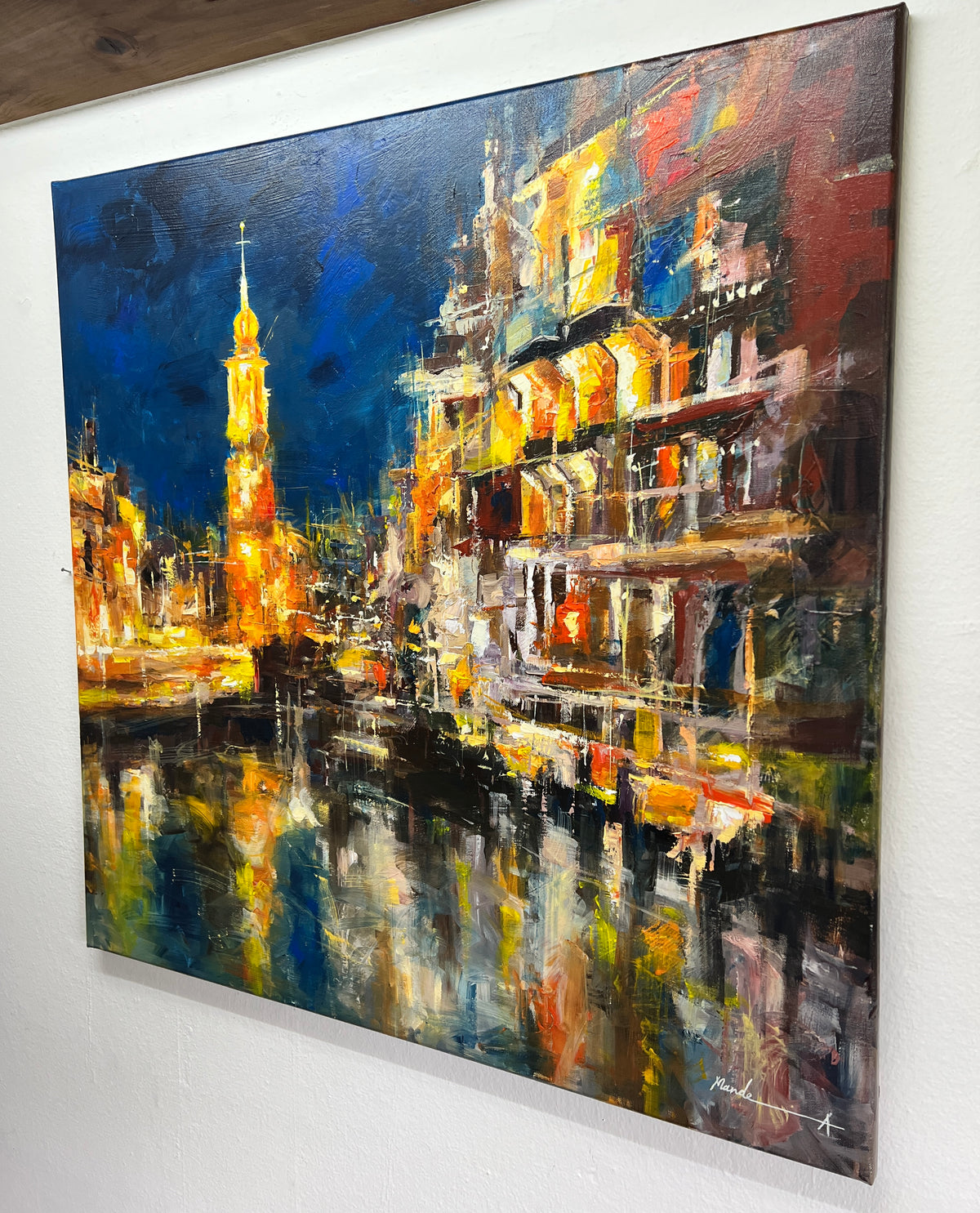 An expressive amsterdam painting of a canal-side city scene at night, mounted on a white wall. The artwork features glowing yellow and orange buildings, a lit tower, and reflections in the water, rendered with thick impasto brushstrokes in deep blue, black, white, and warm golden tones.